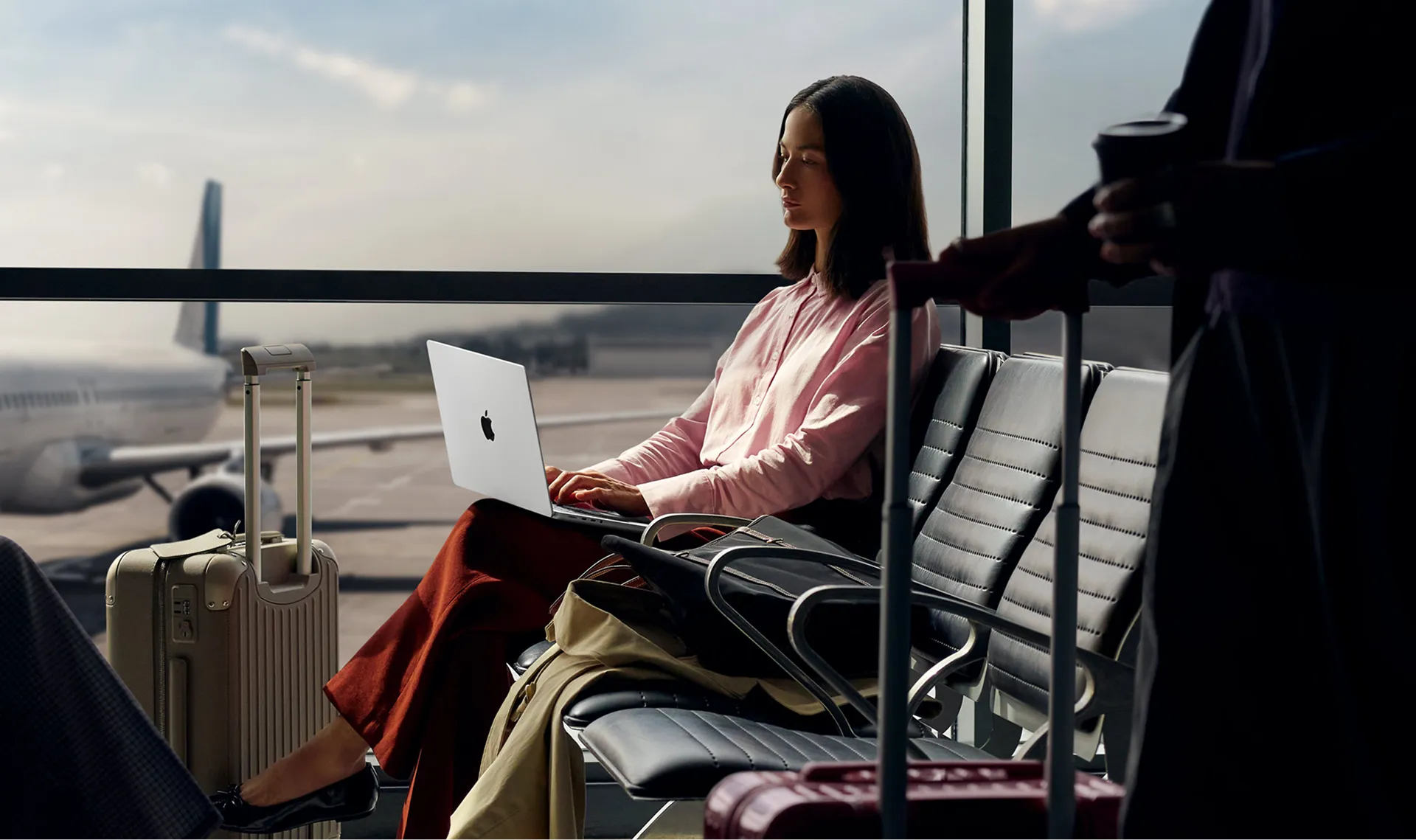 Woman working on a laptop in an airport terminal waiting area with luggage and an airplane visible outside.