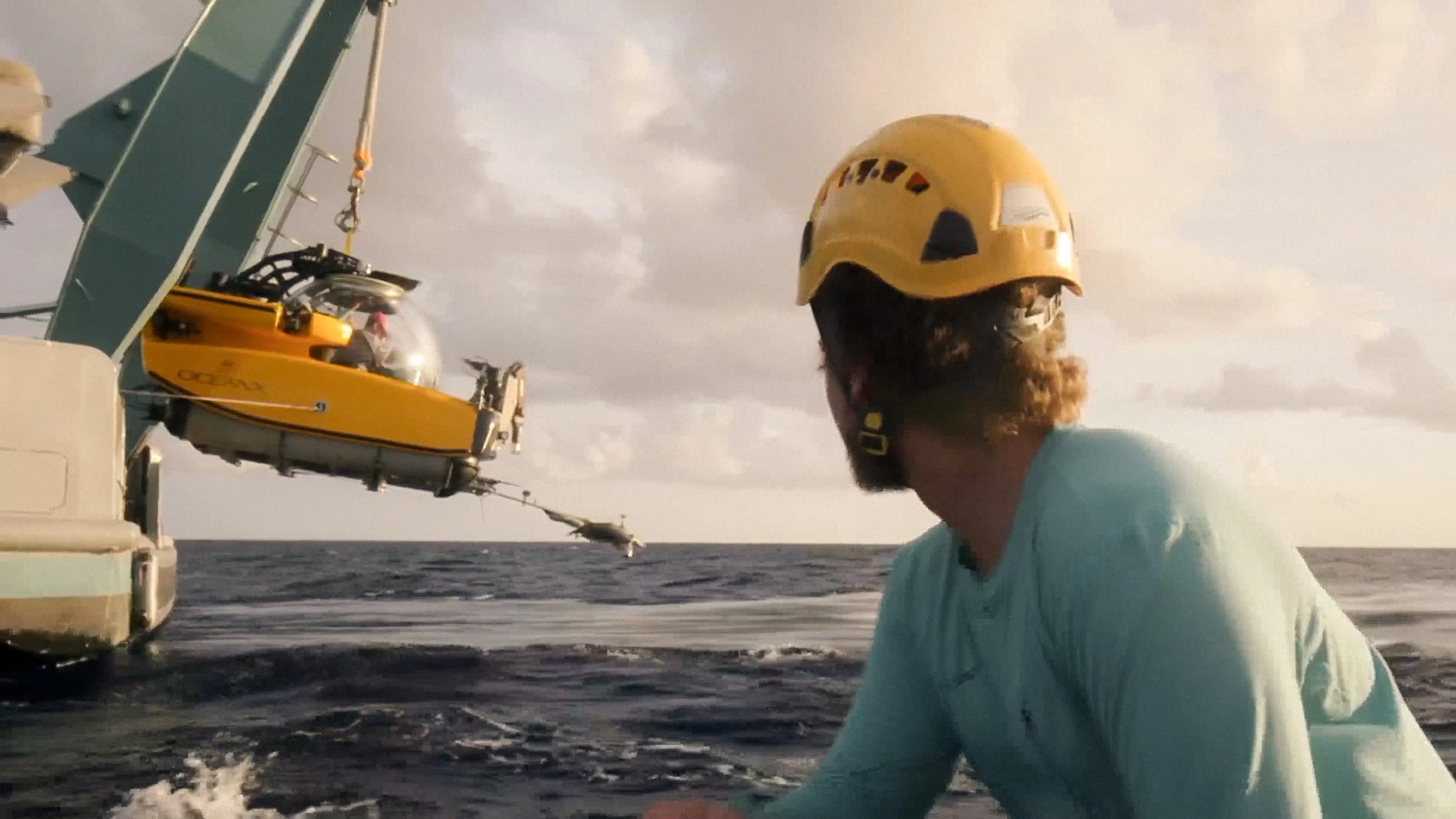 Marine researcher in a hard hat on a boat watching a yellow submarine being lowered into the ocean.