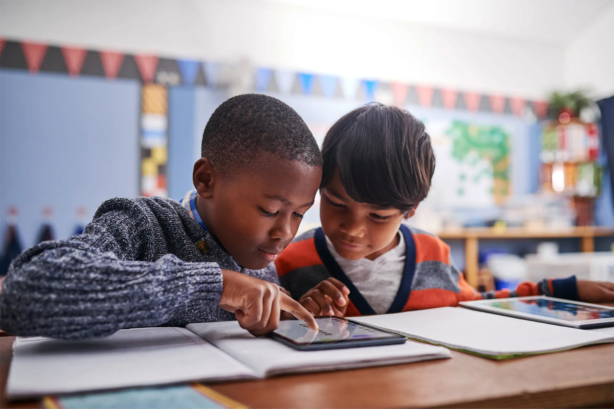 Two young students in a colorful classroom collaborating on a project using an iPad and Apple Pencil.